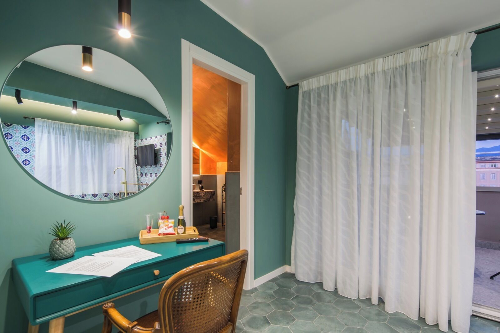 Una habitación con un escritorio color turquesa, una silla, un espejo, una ventana con cortinas blancas y vistas al exterior.