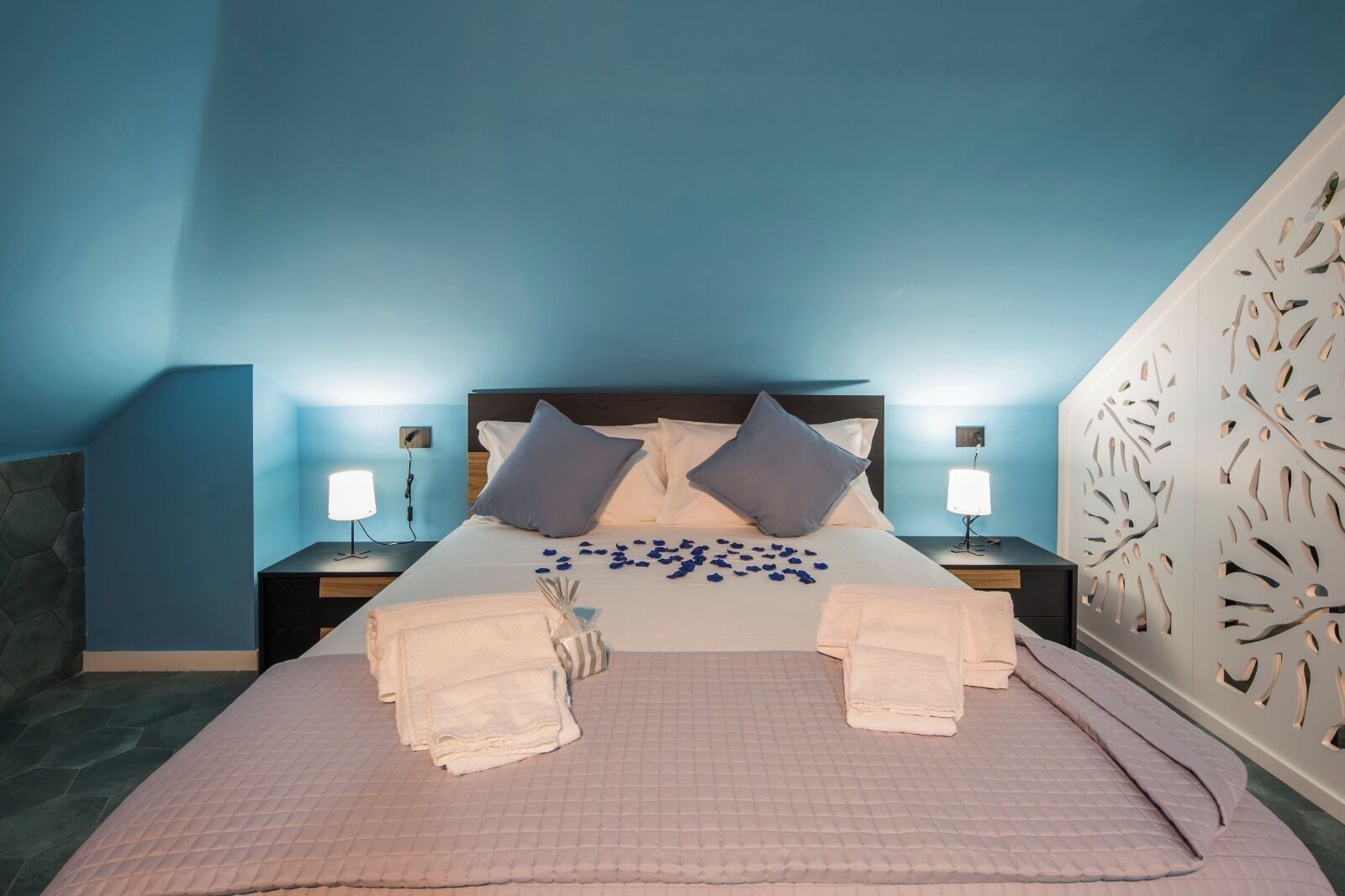 Habitación de hotel con cama, dos mesitas de noche, un panel decorativo en la pared y cabecero.