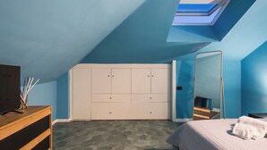 Un dormitorio con techo inclinado, una cama con manta blanca, un aparador de madera, un televisor y un armario con varias puertas.