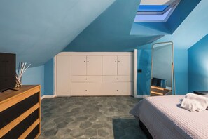 Un dormitorio con techo inclinado, una cama con manta blanca, un aparador de madera, un televisor y un armario con varias puertas.