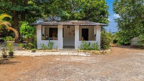 Un piccolo edificio a un piano con un portico, circondato da un vialetto sterrato e da una vegetazione rigogliosa.