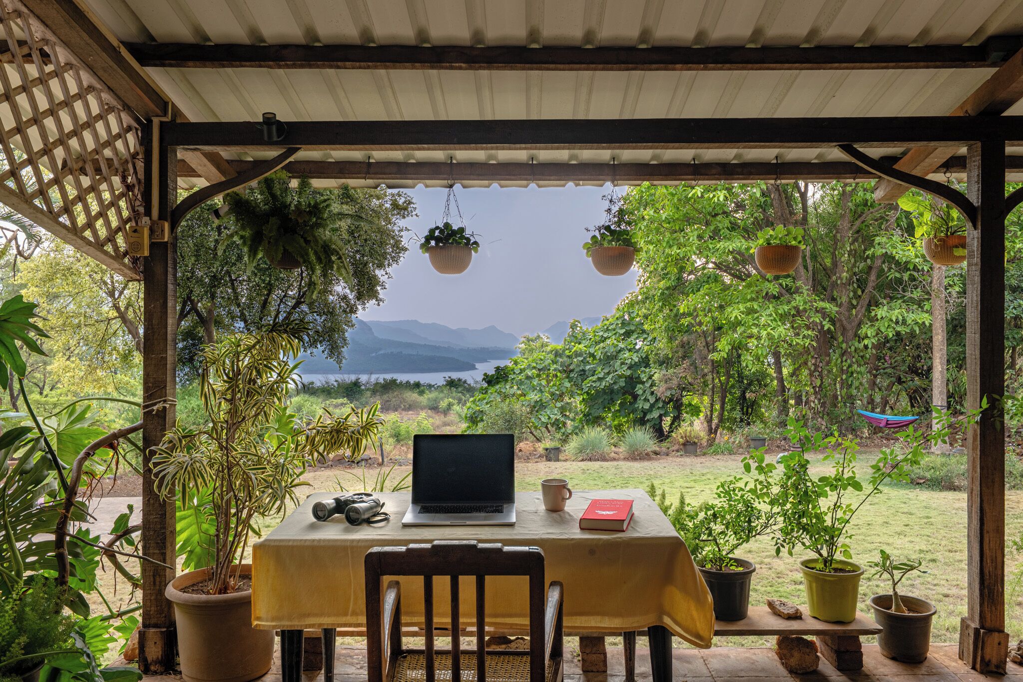 A covered outdoor seating area with a laptop, binoculars, and a book on a table, surrounded by potted plants and overlooking a scenic landscape.