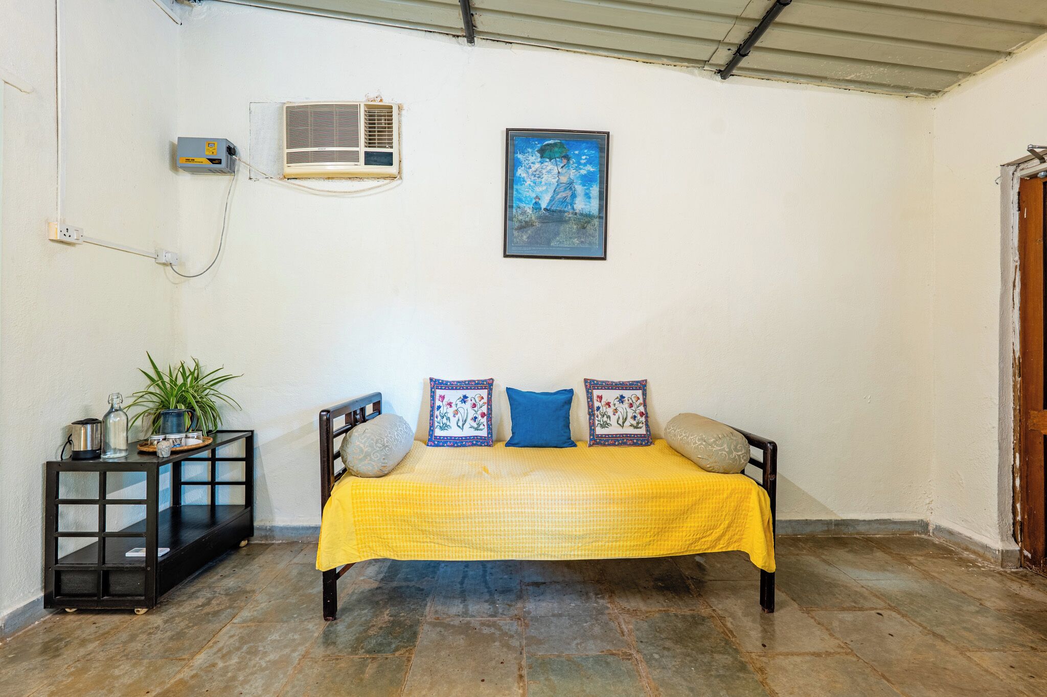 A room with a bed, a small table with a plant, an air conditioning unit, and a framed picture on the wall.