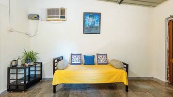 A room with a bed, a small table with a plant, an air conditioning unit, and a framed picture on the wall.