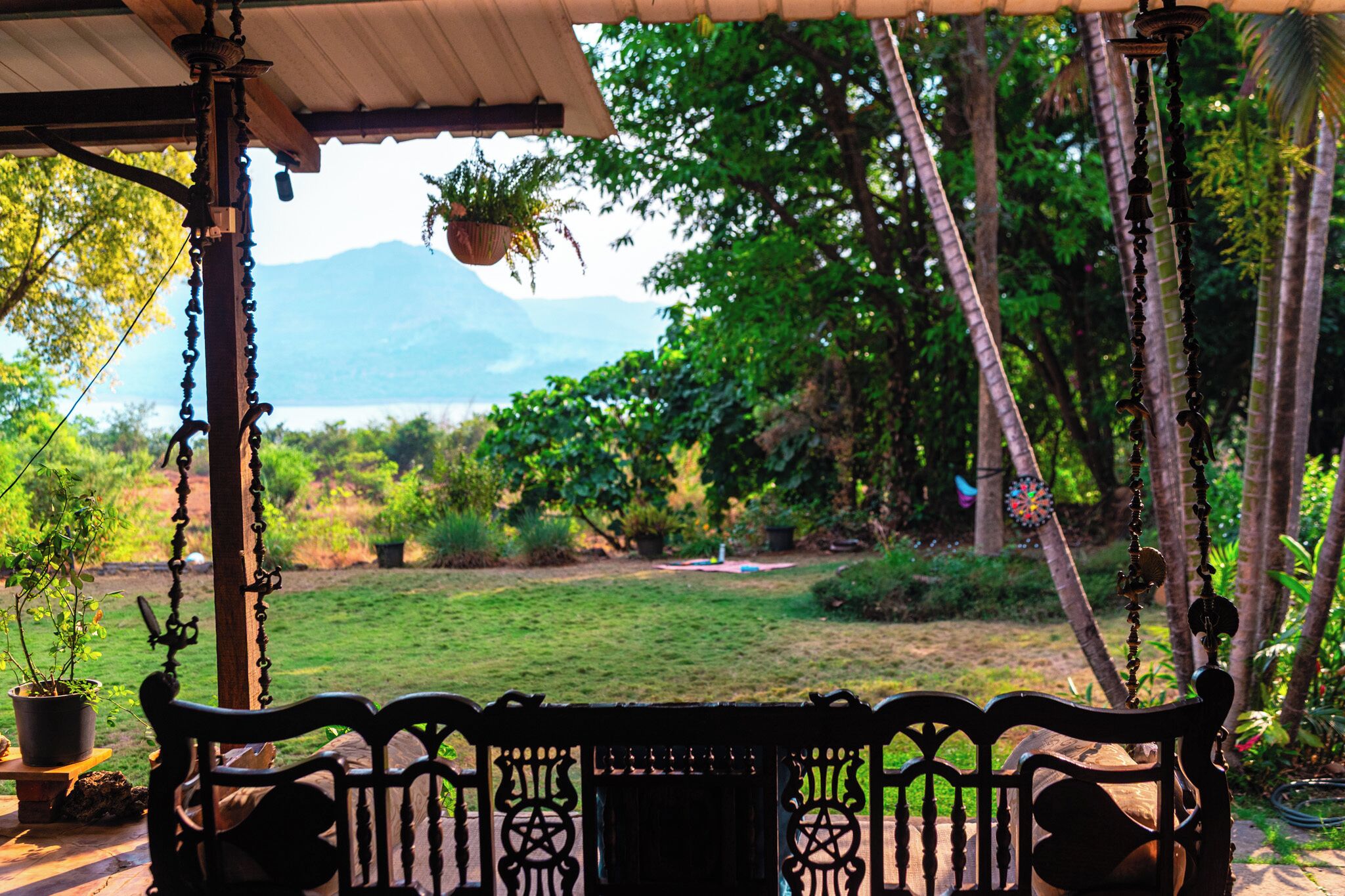 A wooden bench with intricate carvings, a hanging plant, and a view of a lush garden with a mountain in the distance.