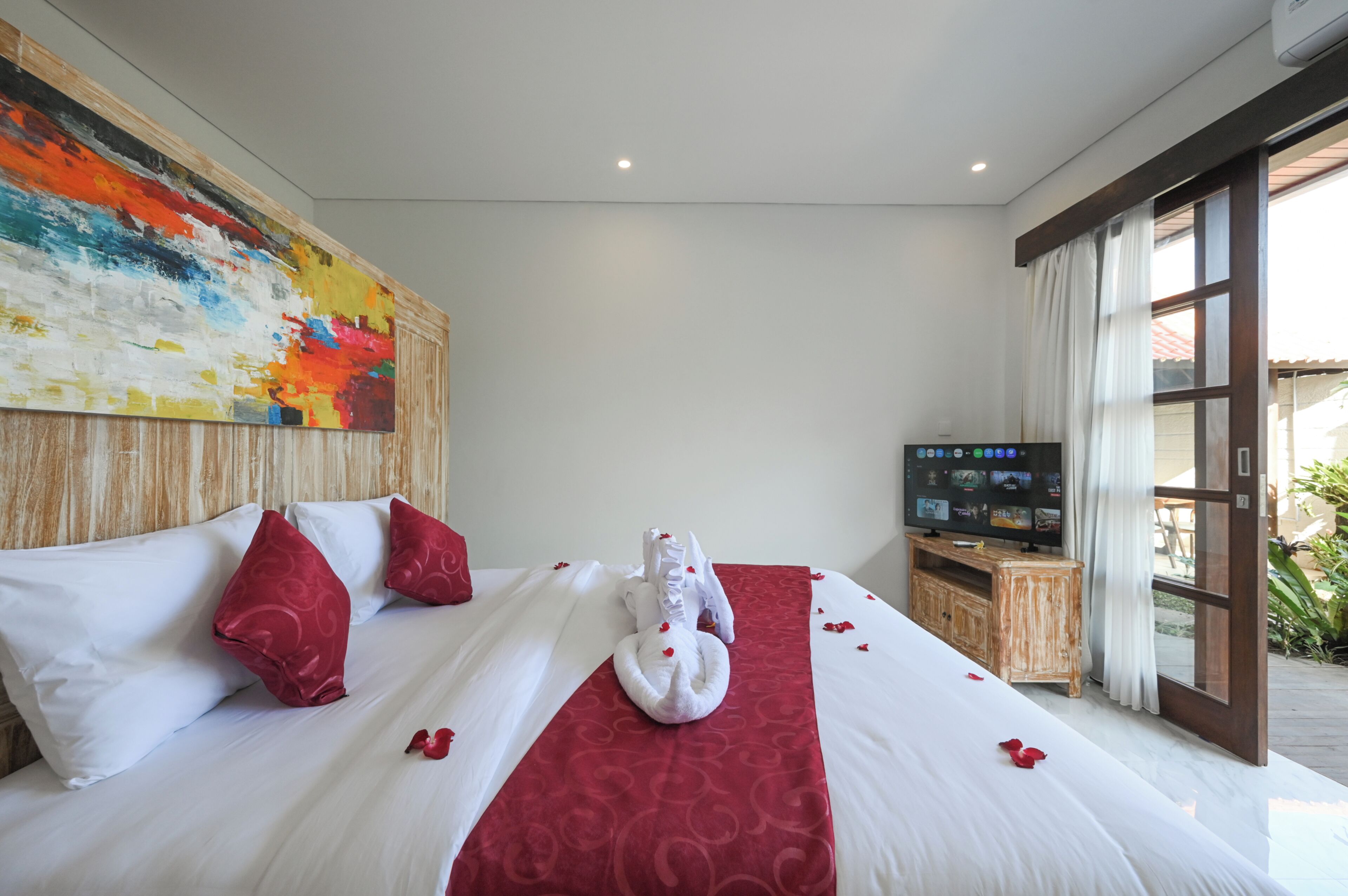 Una habitación de hotel con cama, almohadas, un objeto decorativo rojo, un televisor y un ventanal grande.