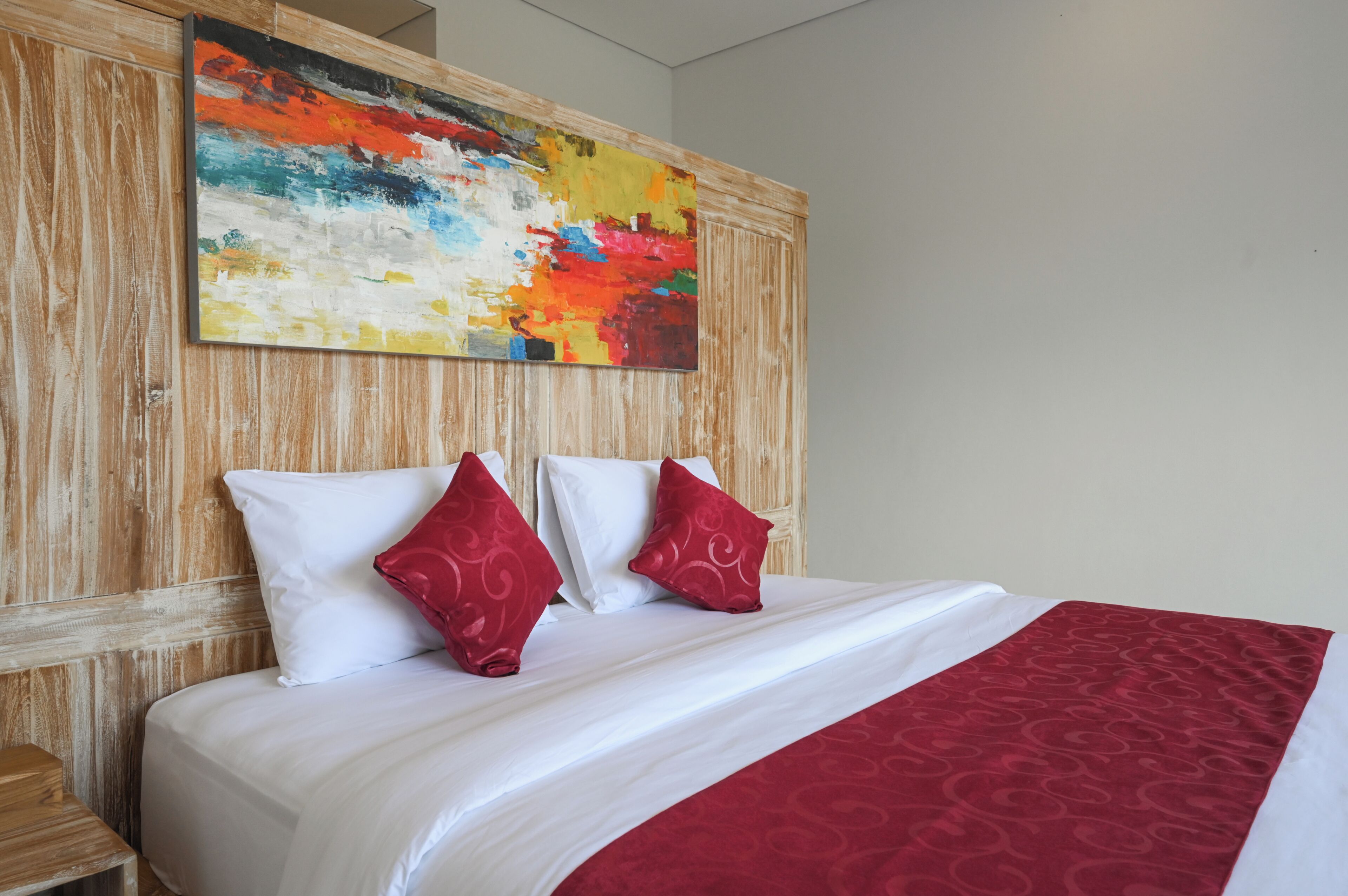 A hotel room with a large bed, white bedding, two red pillows, and a colorful abstract painting on the wooden headboard.