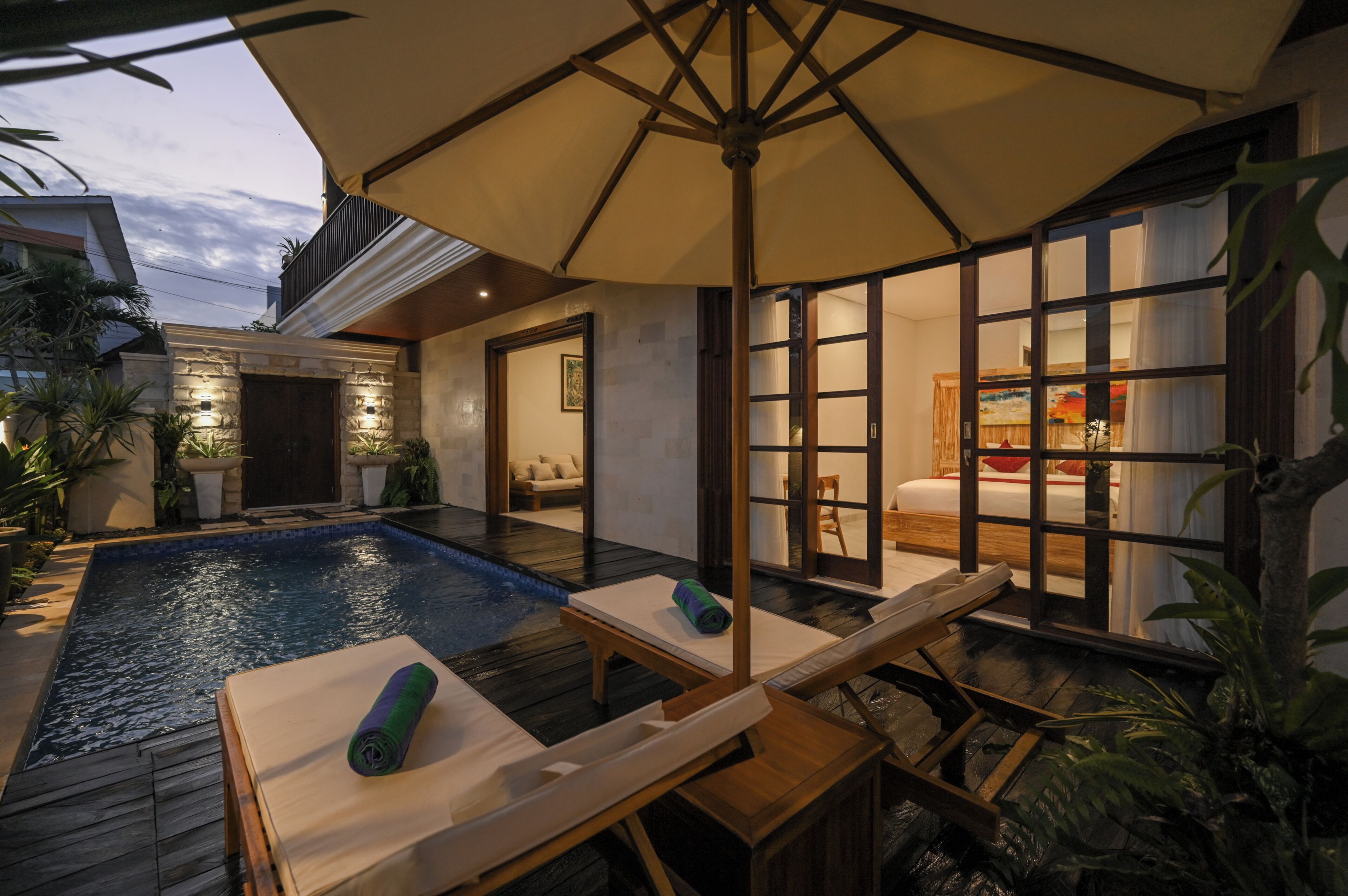 Un área de piscina con sillones de descanso y una sombrilla, una terraza de madera y vista al interior a través de un ventanal.