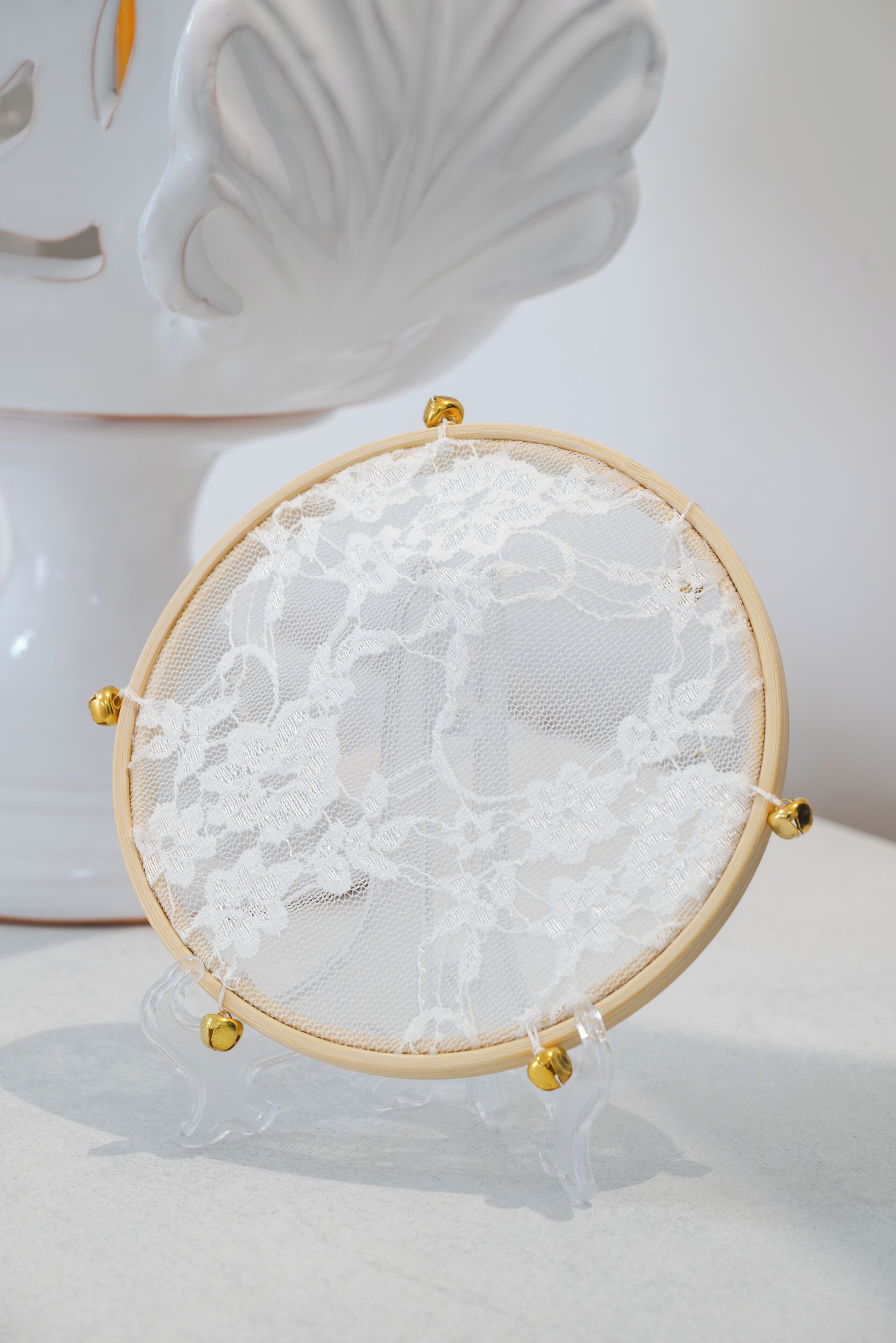 Un aro de bordado redondo de madera con tela de encaje blanco, sostenido por un soporte de plástico transparente con detalles dorados.