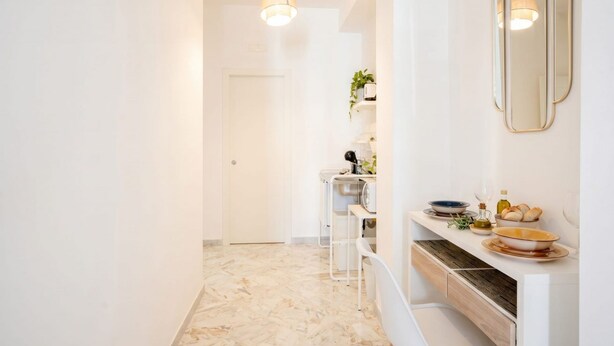 Un pasillo con una puerta blanca, un mostrador blanco con espejo y piso de mármol.