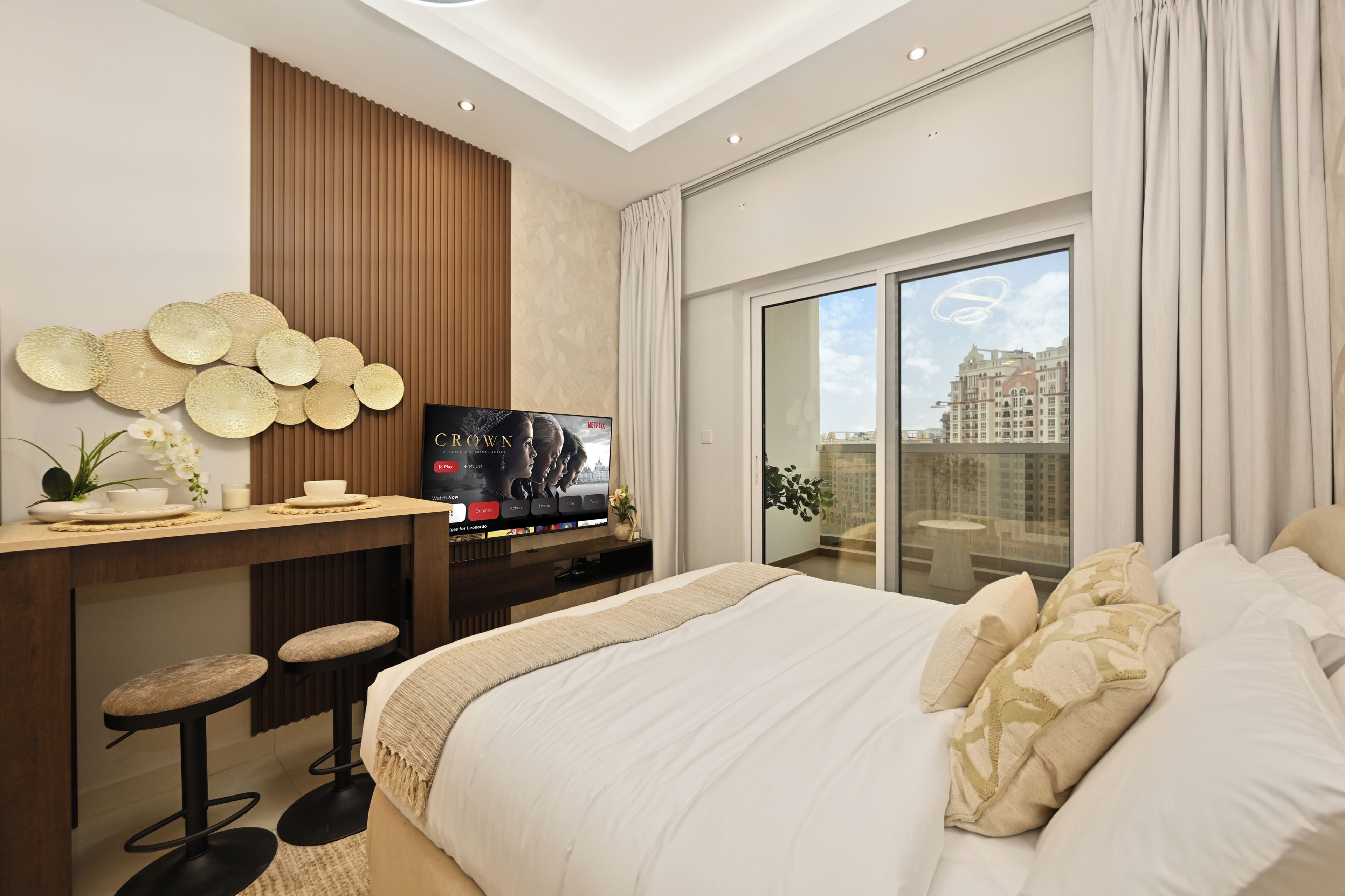 Ein modernes Schlafzimmer mit einem großen Bett, einem Fernseher, einem Schreibtisch mit Hocker und Blick auf eine Stadtlandschaft durch ein großes Fenster.