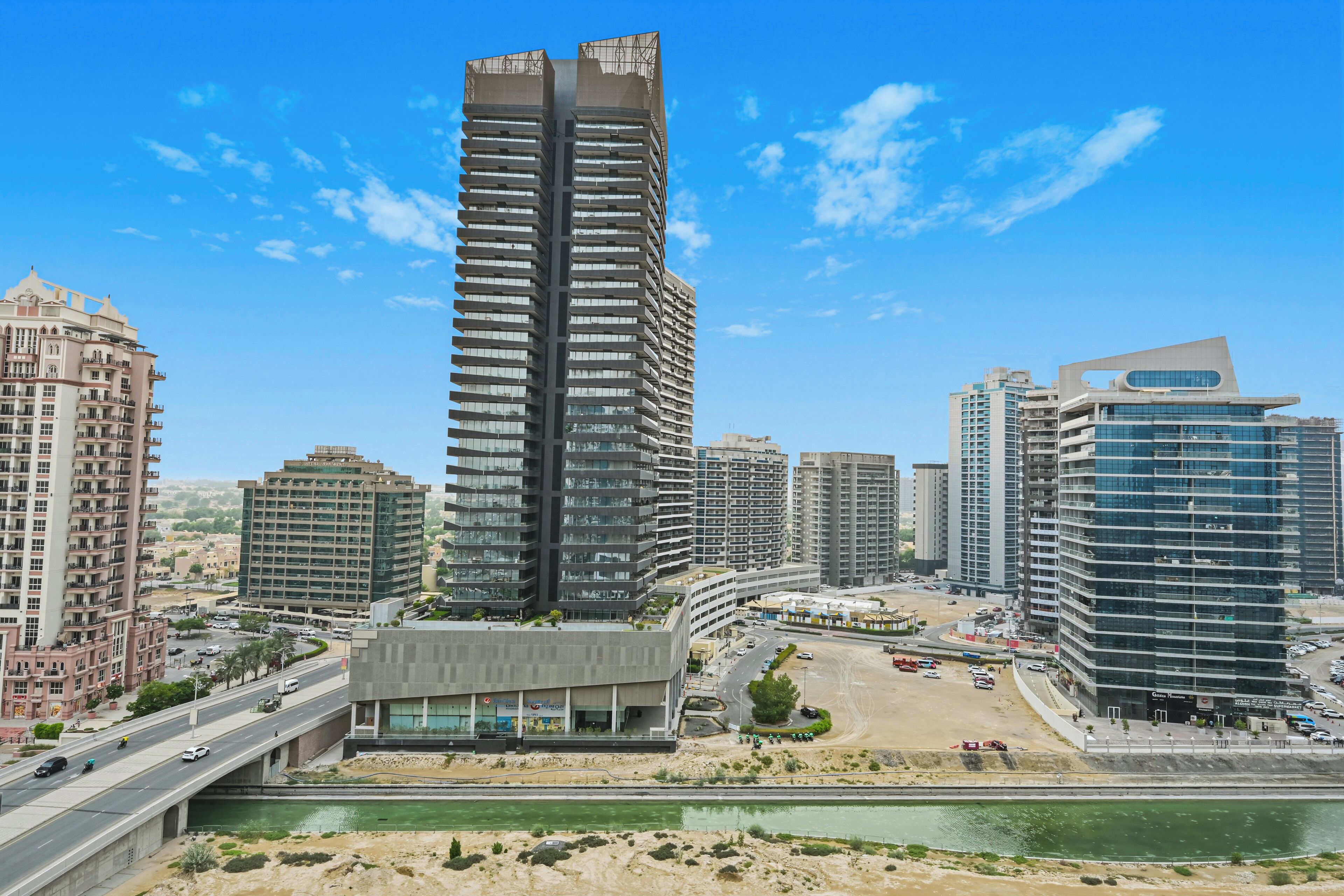 Un immeuble moderne de grande hauteur, doté d’une architecture originale, entouré d’autres bâtiments contemporains et bordé par une rue très fréquentée où circulent de nombreux véhicules.