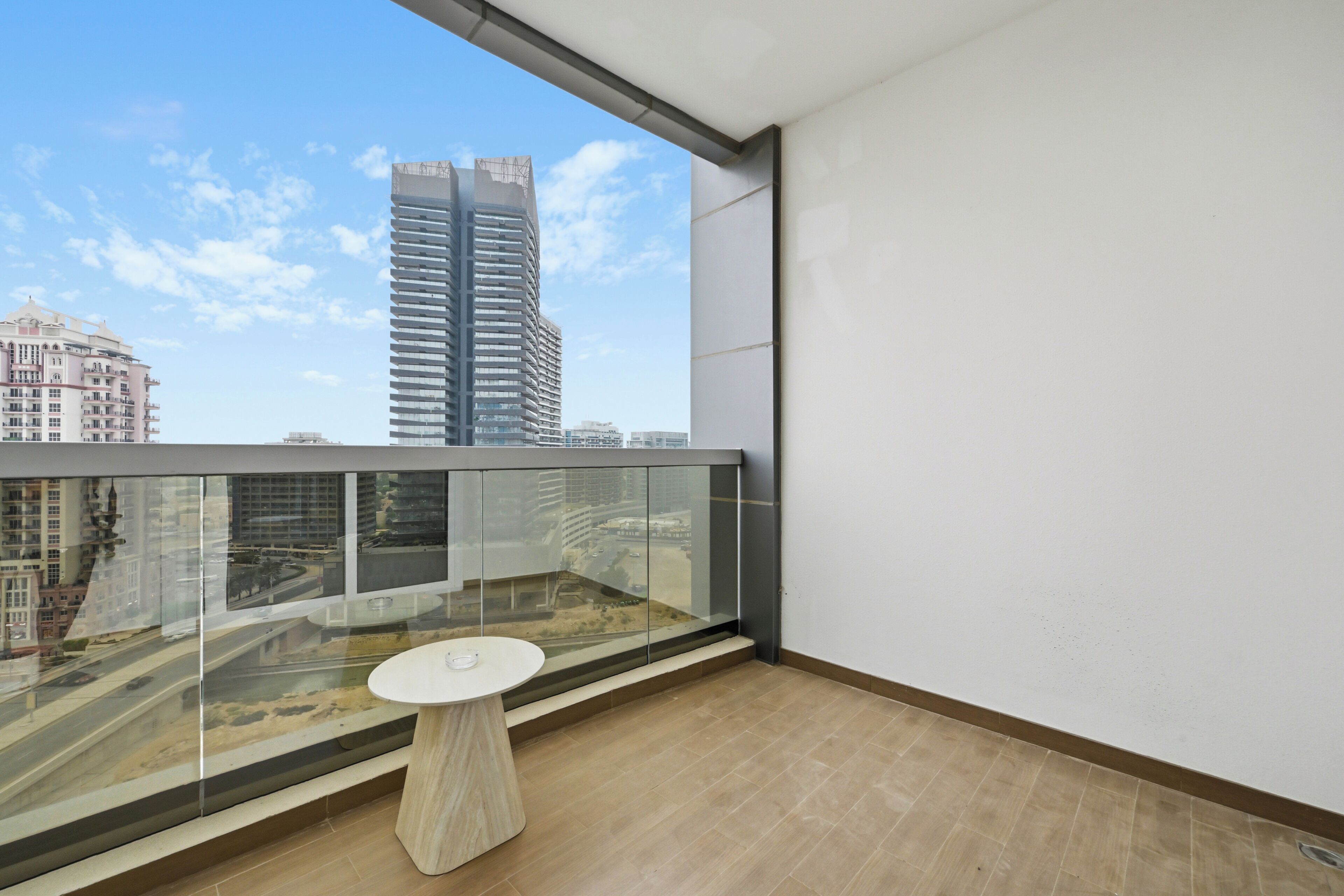 Un balcón con barandilla de vidrio, una pequeña mesa redonda y vistas a edificios de la ciudad.