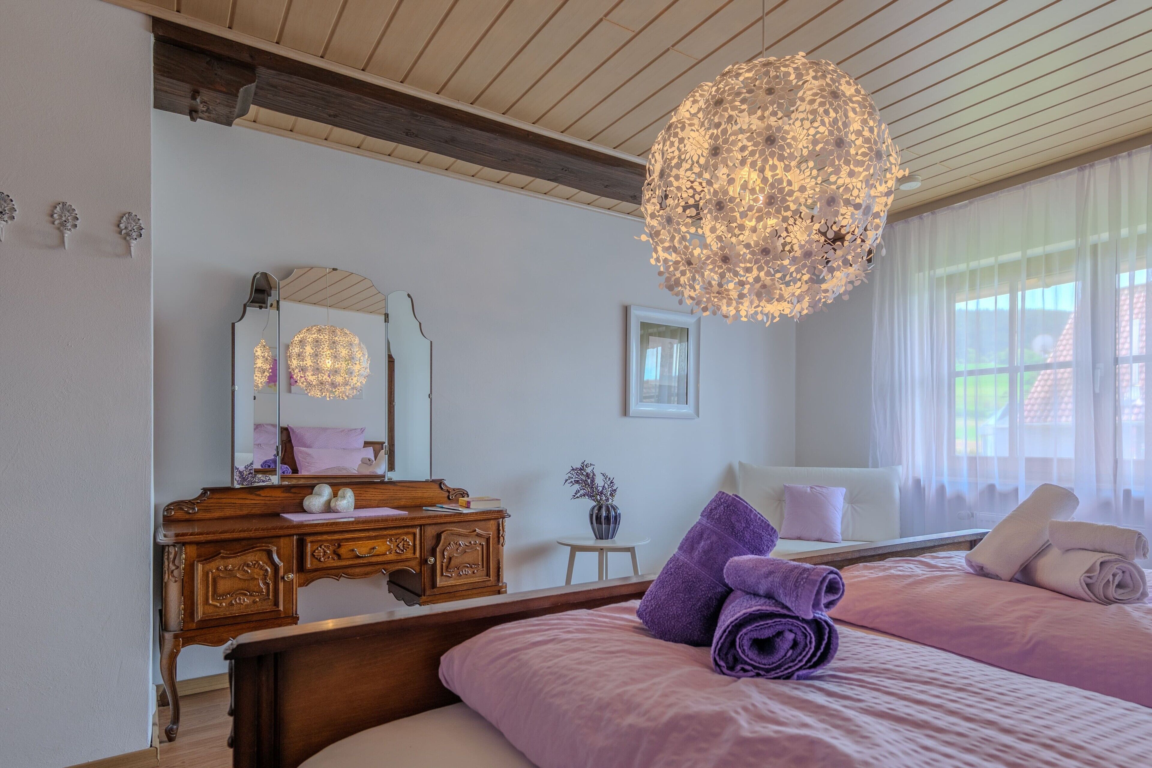 Un dormitorio con cómoda de madera, un espejo grande, una cama con ropa de cama color púrpura y una lámpara de techo esférica original.