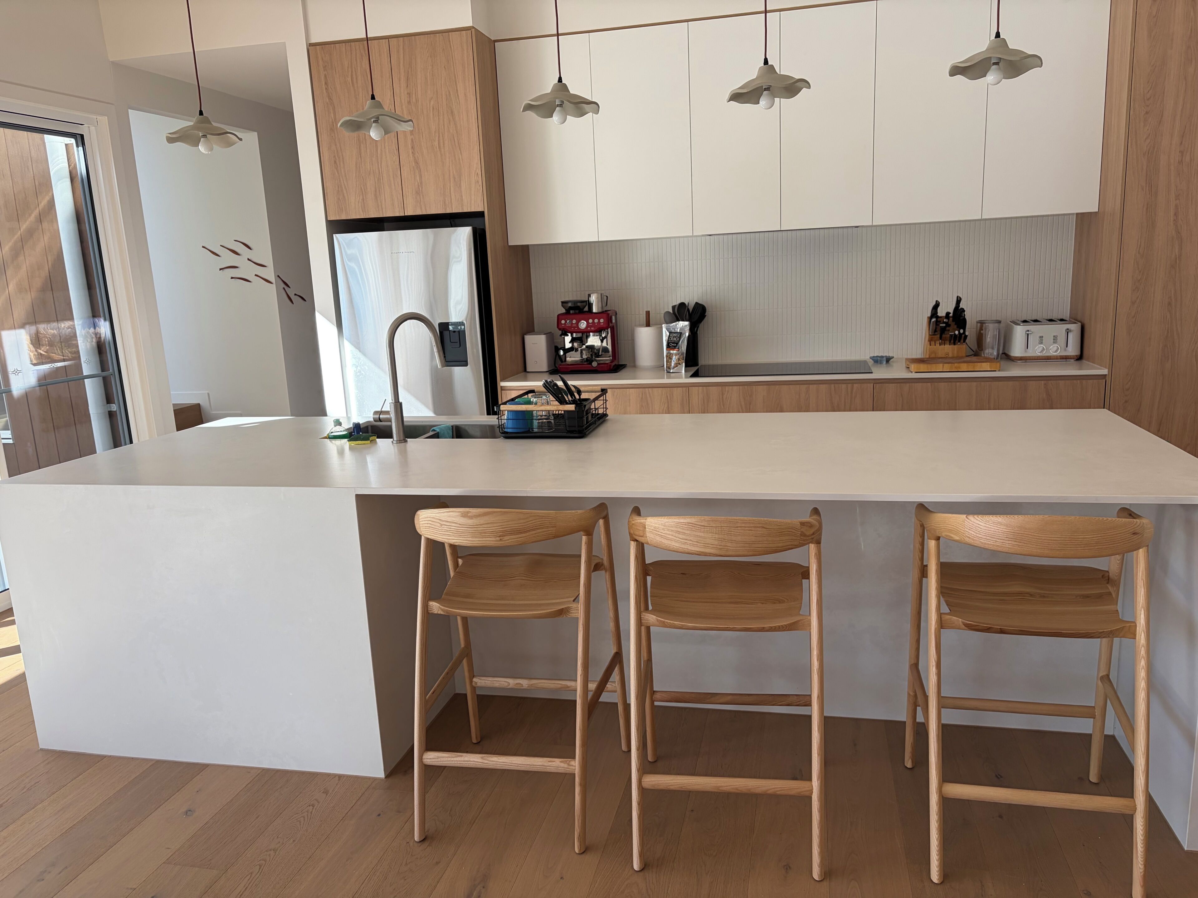 A modern kitchen with a central island, wooden bar stools, pendant lighting, and a stainless steel refrigerator.