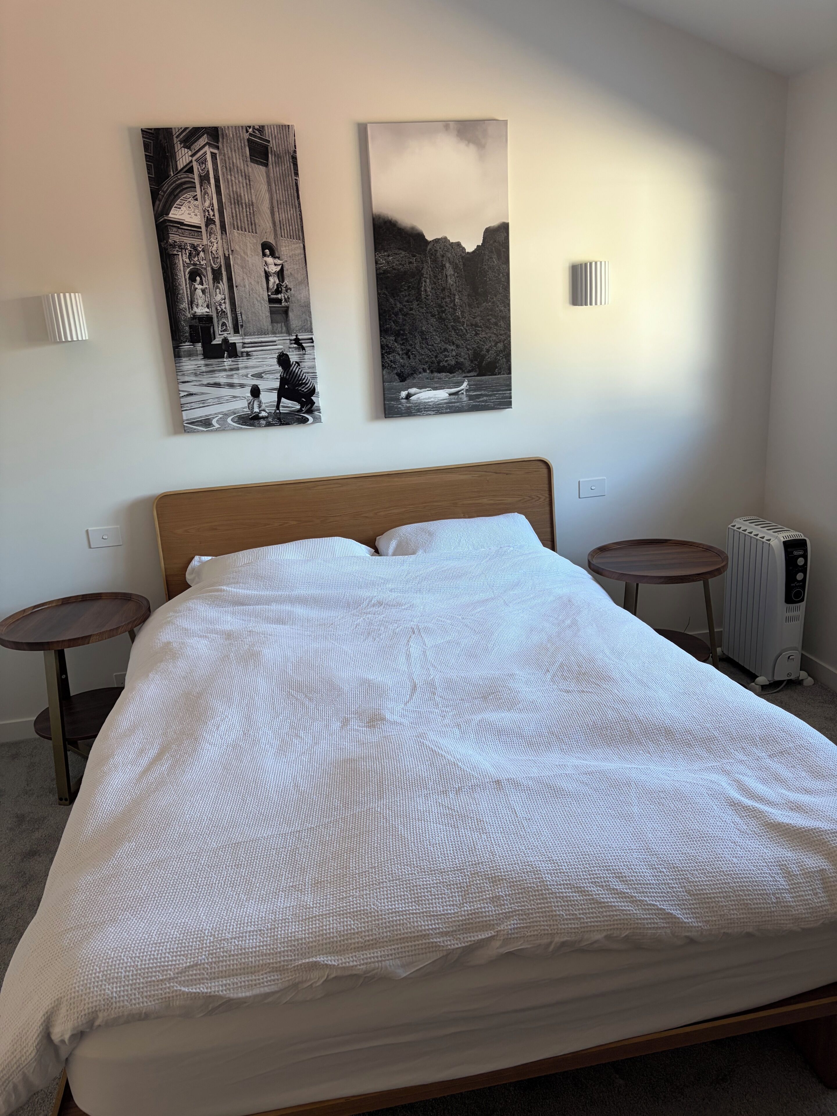 A neatly made bed with a white textured bedspread, two wall-mounted picture frames, a round bedside table, and a radiator.