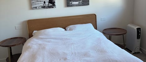 Ein ordentlich bezogenes Bett mit weißer, strukturierter Bettdecke, zwei an der Wand befestigte Bilderrahmen, ein runder Nachttisch und ein Heizkörper.