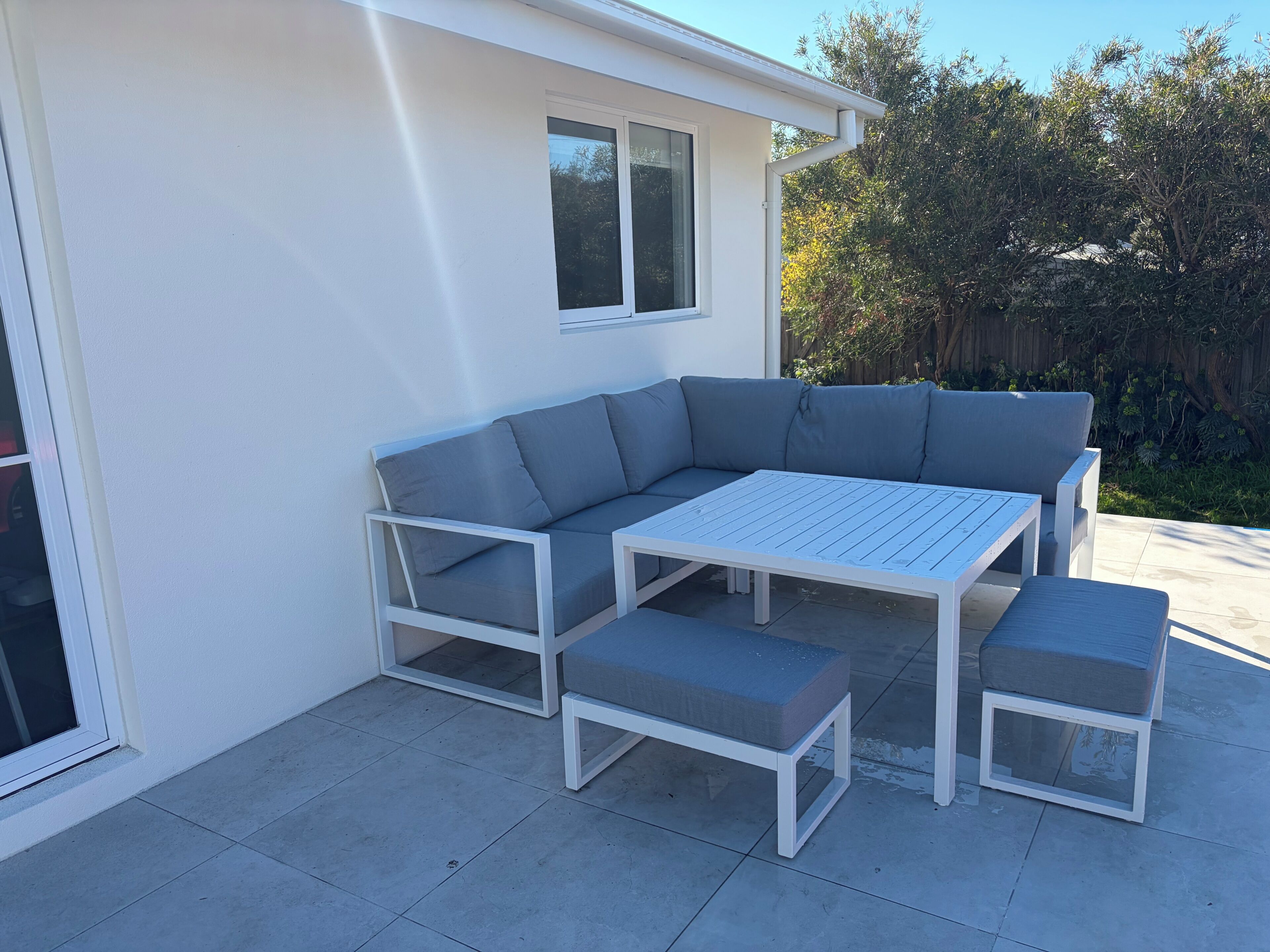 Un patio extérieur moderne avec un canapé d’angle gris, une table blanche et des poufs assortis.