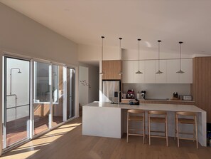 A modern kitchen with a large island, bar stools, pendant lighting, and a sliding glass door leading to a balcony. - Tiberius House | Brand New Luxury Retreat with Heated Pool, Sauna & Pickleball (Saint Andrews Beach)