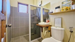 Un baño con inodoro, lavamanos, cabina de ducha y ventana con persianas.
