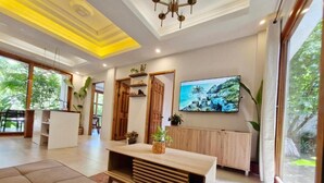 Una sala moderna con un sofá, una mesa de centro de madera, un televisor montado en la pared y un ventanal con vistas a la vegetación.