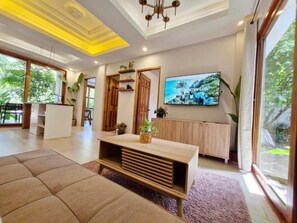 A modern living room with a sofa, a wooden coffee table, a TV mounted on the wall, and a large window with a view of greenery. - Sing Siargao (General Luna)