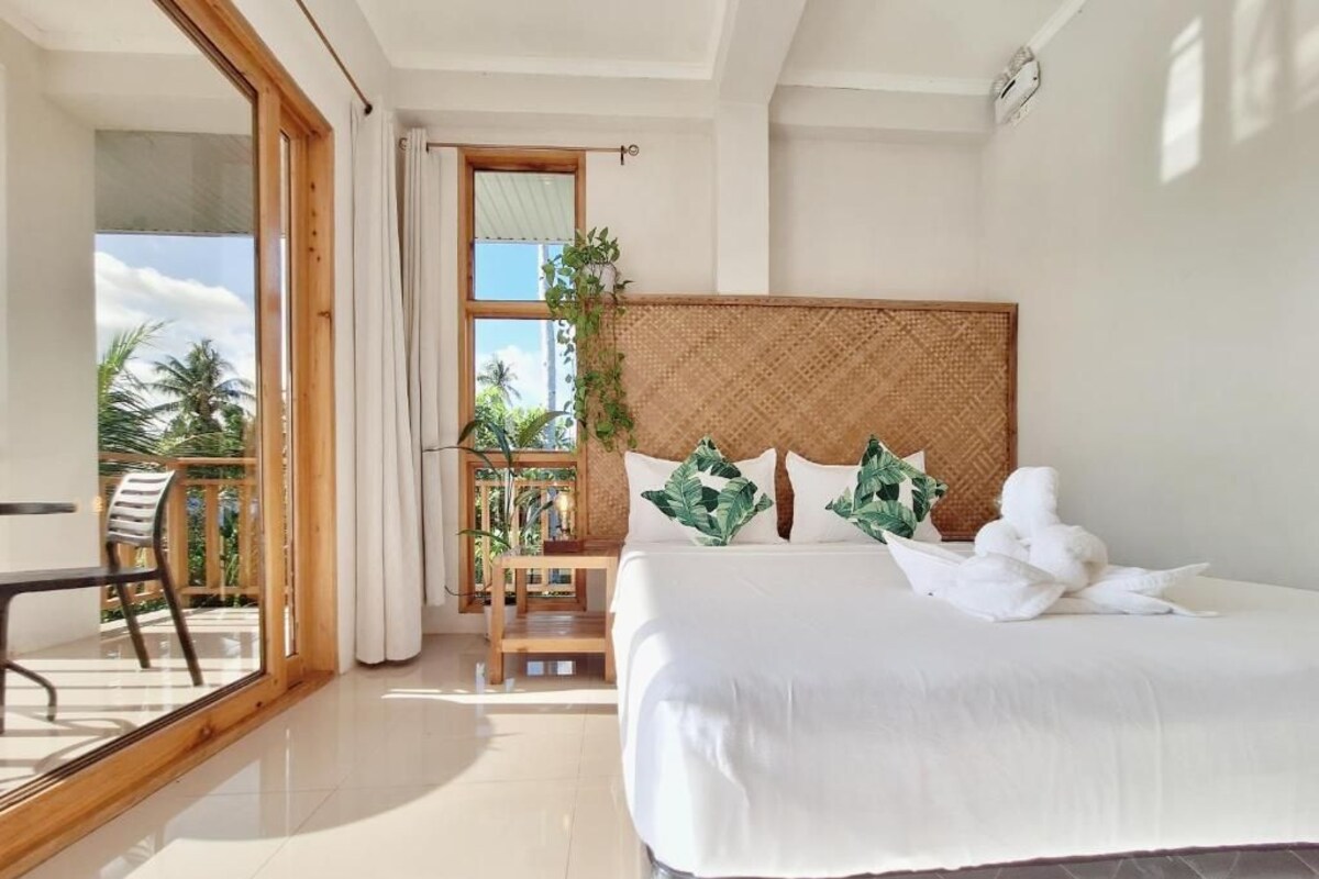 A hotel room with a bed, a chair, a table, a balcony with a view of palm trees, and a window with curtains.