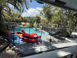 Piscina com toboáguas infláveis e espreguiçadeiras, cercada por palmeiras e um espaço delimitado por cerca.