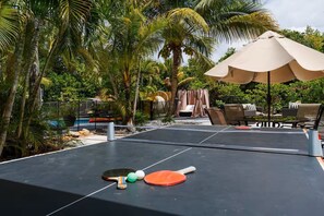 A ping pong table with two paddles and three balls, surrounded by outdoor furniture and palm trees.