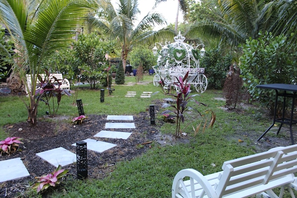 Um jardim com um banco branco, uma estrutura branca decorativa e um caminho pavimentado.