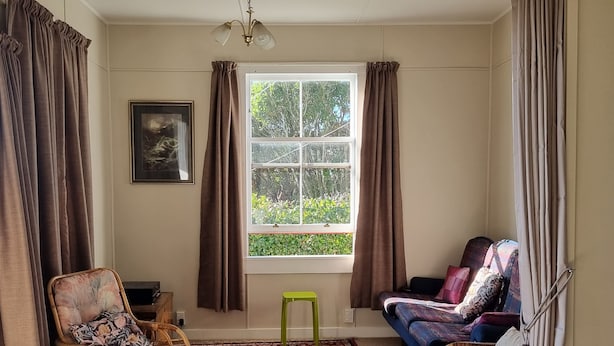 A room with a large window, a wicker chair, a sofa, and a small stool.