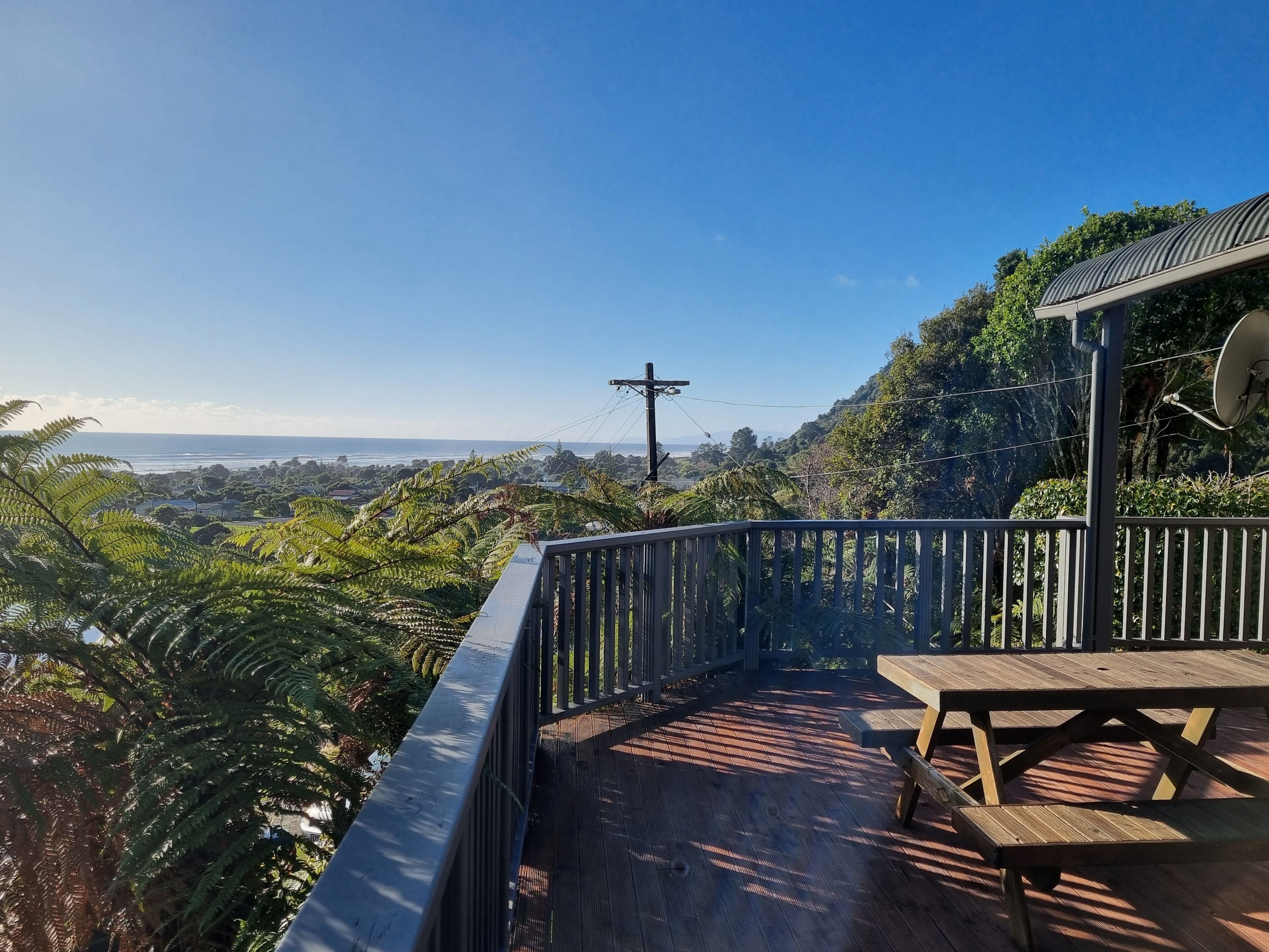 A wooden deck with a picnic table, overlooking a coastal landscape with dense greenery and a clear sky.