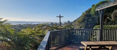 A wooden deck with a picnic table, overlooking a coastal landscape with dense greenery and a clear sky.