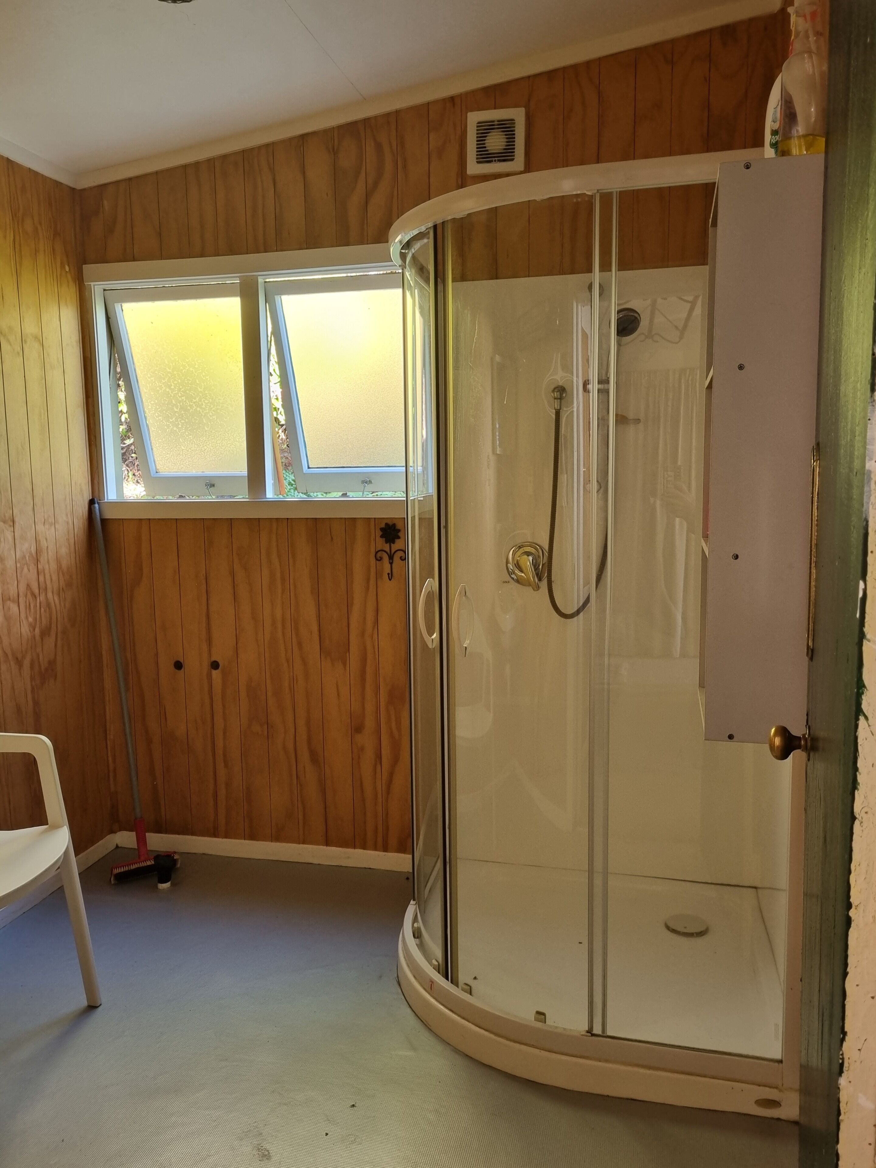 A room with a glass-enclosed shower, wooden walls, a window, and a white chair.