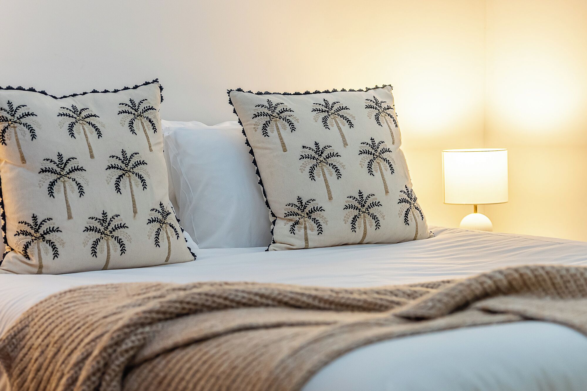 A bed with two pillows featuring palm tree patterns, a bedside lamp, and a knitted throw blanket.