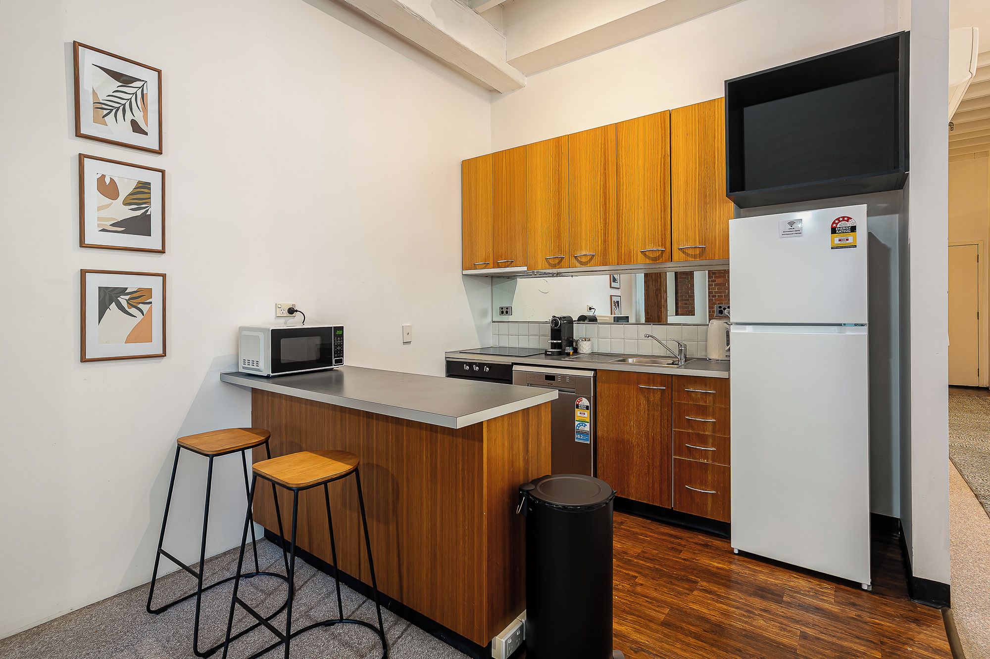 A compact kitchen with wooden cabinets, a microwave, a refrigerator, and a small bar stool.