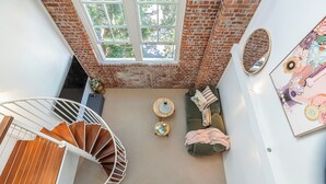 A loft-style room with a spiral staircase, a green sofa set, a round wooden coffee table, and a large window with a view of trees. - Mactaggarts Place Apartments (Teneriffe)