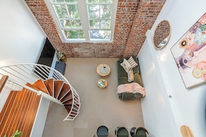 A loft-style room with a spiral staircase, a green sofa set, a round wooden coffee table, and a large window with a view of trees. - Mactaggarts Place Apartments (Teneriffe)