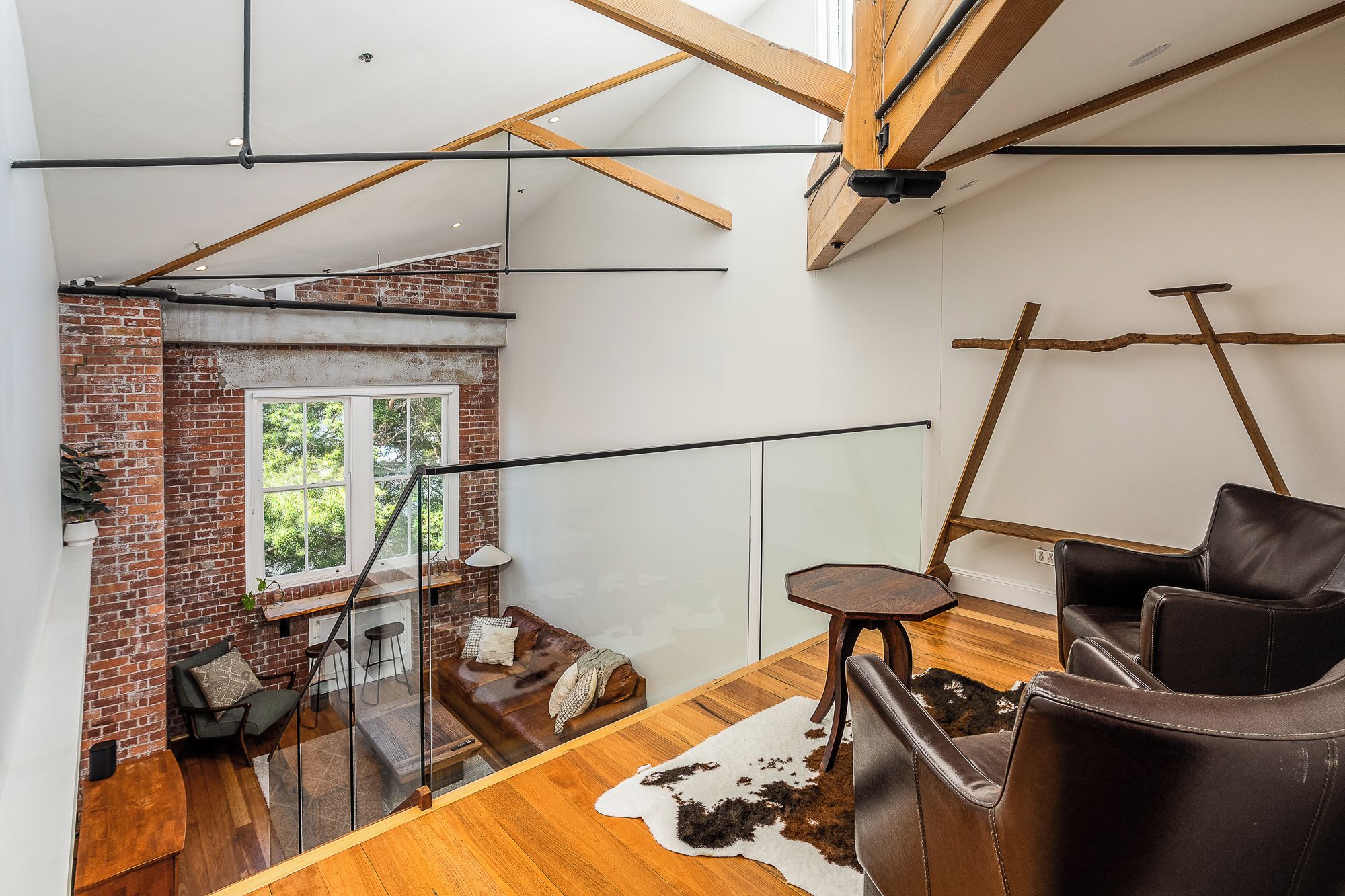 A loft-style room with exposed wooden beams, a brick wall, a glass railing, and a leather sofa.