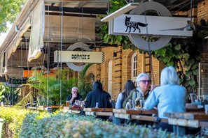 An outdoor seating area with tables and chairs, a hanging sign with a cat logo, and a building with a brick facade in the background. - Mactaggarts Place Apartments (Teneriffe)