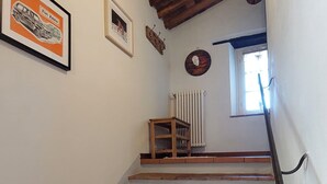 Un escalier avec une rampe en bois, des murs blancs et un sol en briques menant à un palier où se trouvent un radiateur et une petite table.