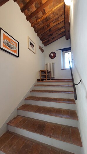 A staircase with a wooden handrail, white walls, and a brick floor leading to a landing with a radiator and a small table. - In Celli, Between the Woods and the Sky - Apartment in an Old Farmhouse Surrounded by Greenery (San Martino in Freddana - Monsagrati)
