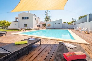 A swimming pool with lounge chairs and a shaded area, surrounded by a modern building.
