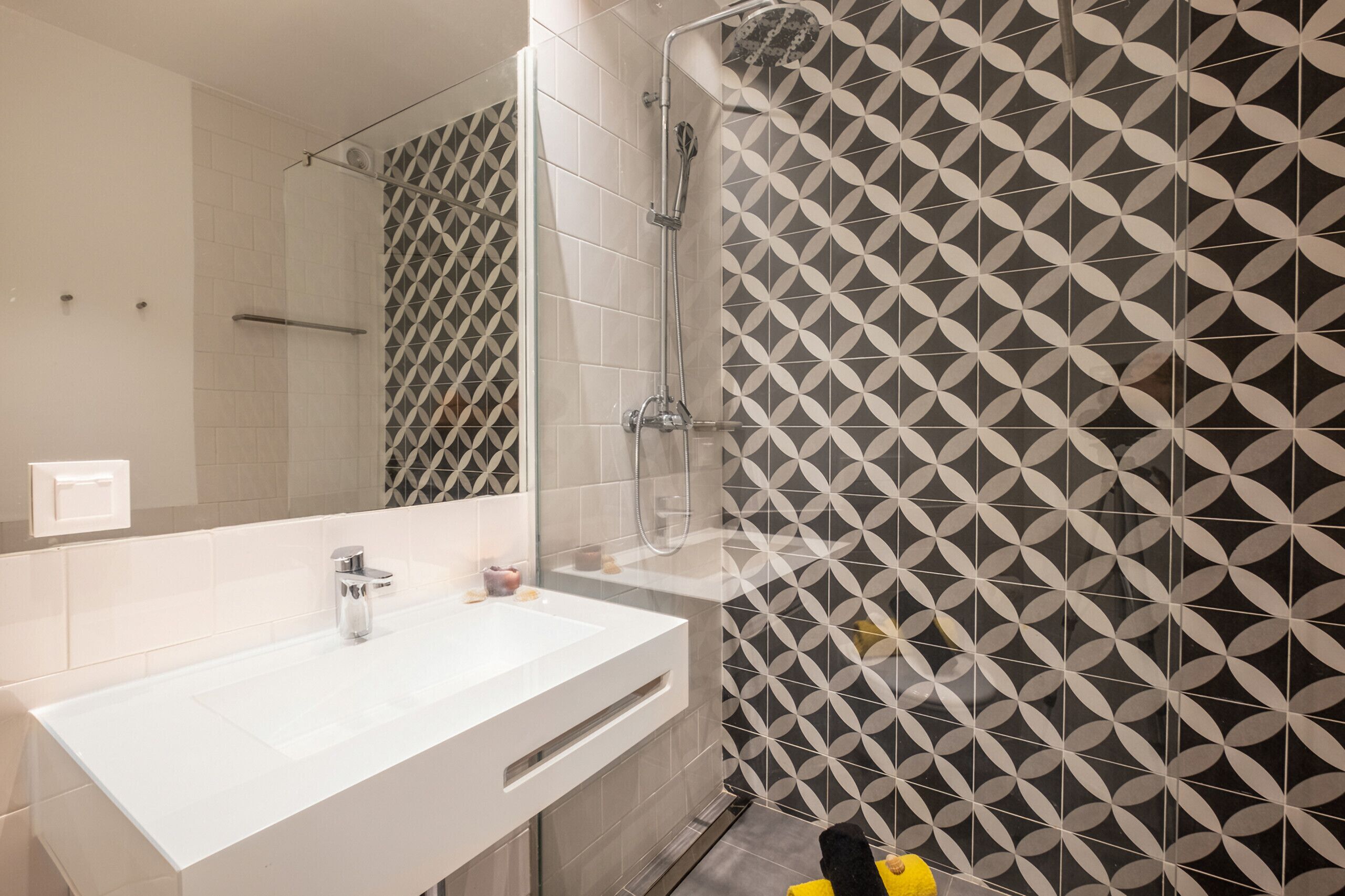 A modern bathroom with a white sink, a shower with a geometric tile pattern, and a mirror.