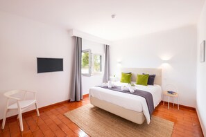 A bedroom with a bed, a chair, a TV, a window, and a bedside table.