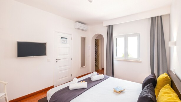 A hotel room with a bed, a flat-screen TV, a door, a window with curtains, and a small table with a book.
