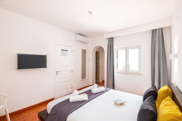 A hotel room with a bed, a flat-screen TV, a door, a window with curtains, and a small table with a book.