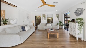 A spacious living room with a wooden floor, a ceiling fan, and a large window offering a view of the outdoors. - Waterfront Retreat in the heart of Mooloolaba (mooloolaba)