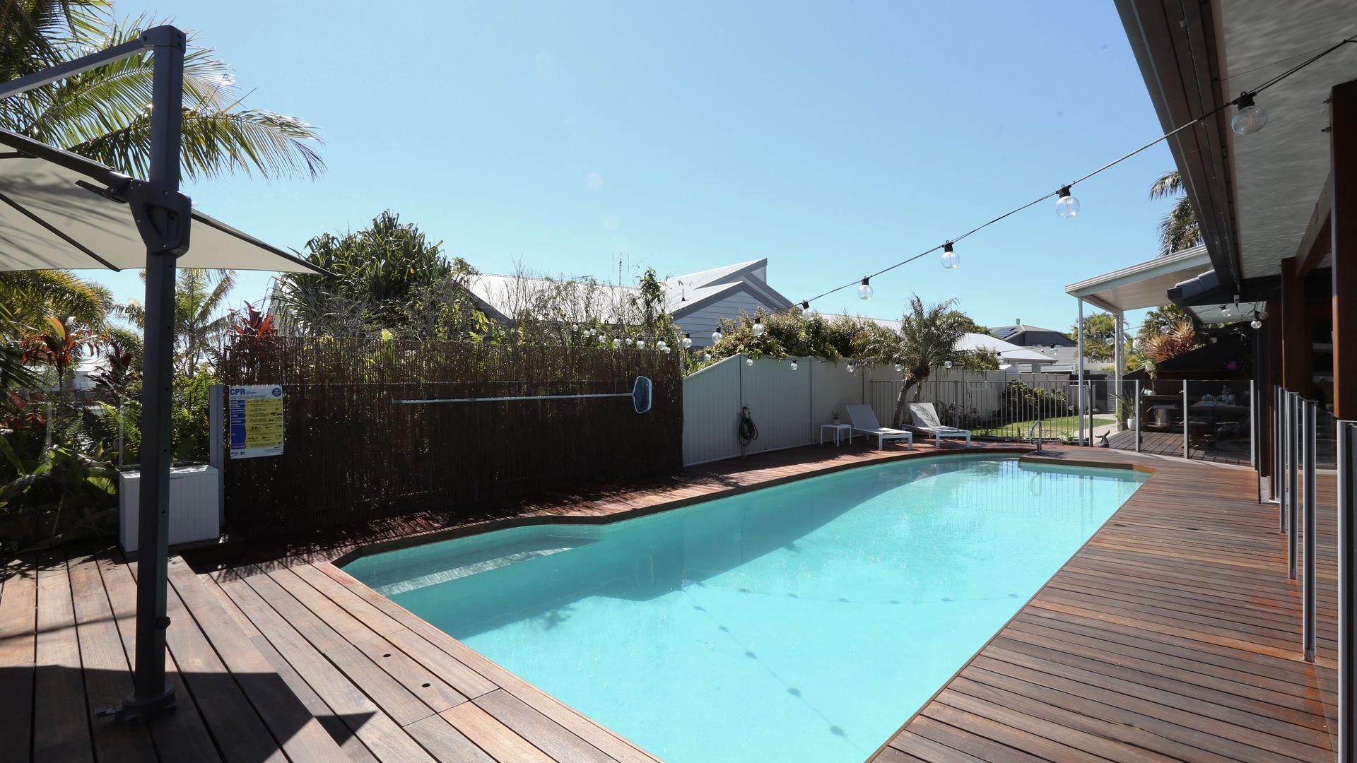 Una piscina con plataforma de madera, zona de estar techada y jardín vallado.