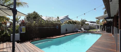 Una piscina con plataforma de madera, zona de estar techada y jardín vallado.
